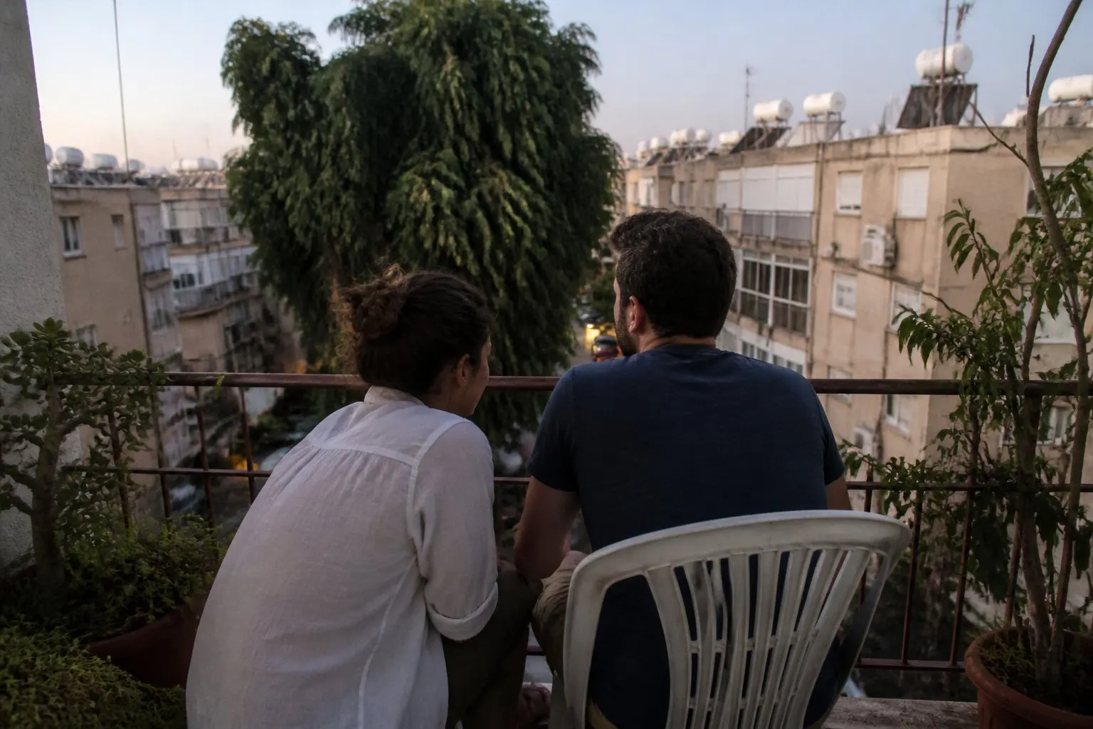 זוג בשנות השלושים יושב במרפסת של דירה בעיר בשעת ערב מוקדמת, מול עץ פיקוס וגגות עם דודי שמש, היא רוכנת לעברו והוא מביט אל הרחוב, מצולמים מאחור